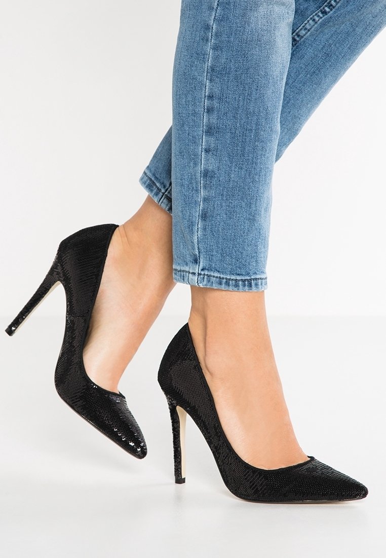 Fötter i svarta paljettprydda spetsiga klackar som är kombinerade med ljusblå avklippta jeans på vit bakgrund.