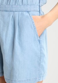 Shorts en chambray bleu clair avec une coupe décontractée, une taille élastique, des poches latérales avec des coutures apparentes et une texture lisse.