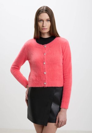 Cardigan fuzzy color corallo chiaro con scollatura rotonda, maniche lunghe e cinque bottoni decorativi. Abbinato a una mini gonna in pelle nera.