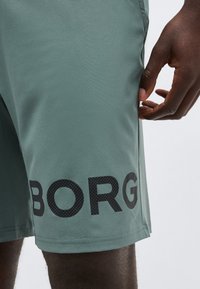 Közeli kép egy személyről, aki világoszöld rövidnadrágot visel, amelynek lábszárán fekete "BORG" felirat található, a kezét a rövidnadrág oldalbélésénél nyugszik.