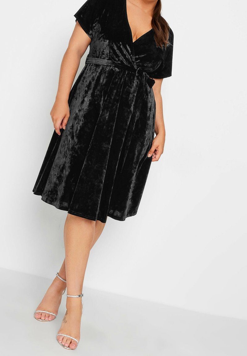 Černé sametové šaty s krátkými rukávy, výstřihem do V a skládací sukní. Stříbrné sandály na kotníku dotvářejí celý outfit.