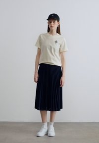 Μπεζ βαμβακερό t-shirt με μικρό τυπωμένο σχέδιο, συνδυασμένο με μπλε πλισέ φούστα. Μοντέλο φοράει μαύρο καπέλο και λευκά αθλητικά παπούτσια.