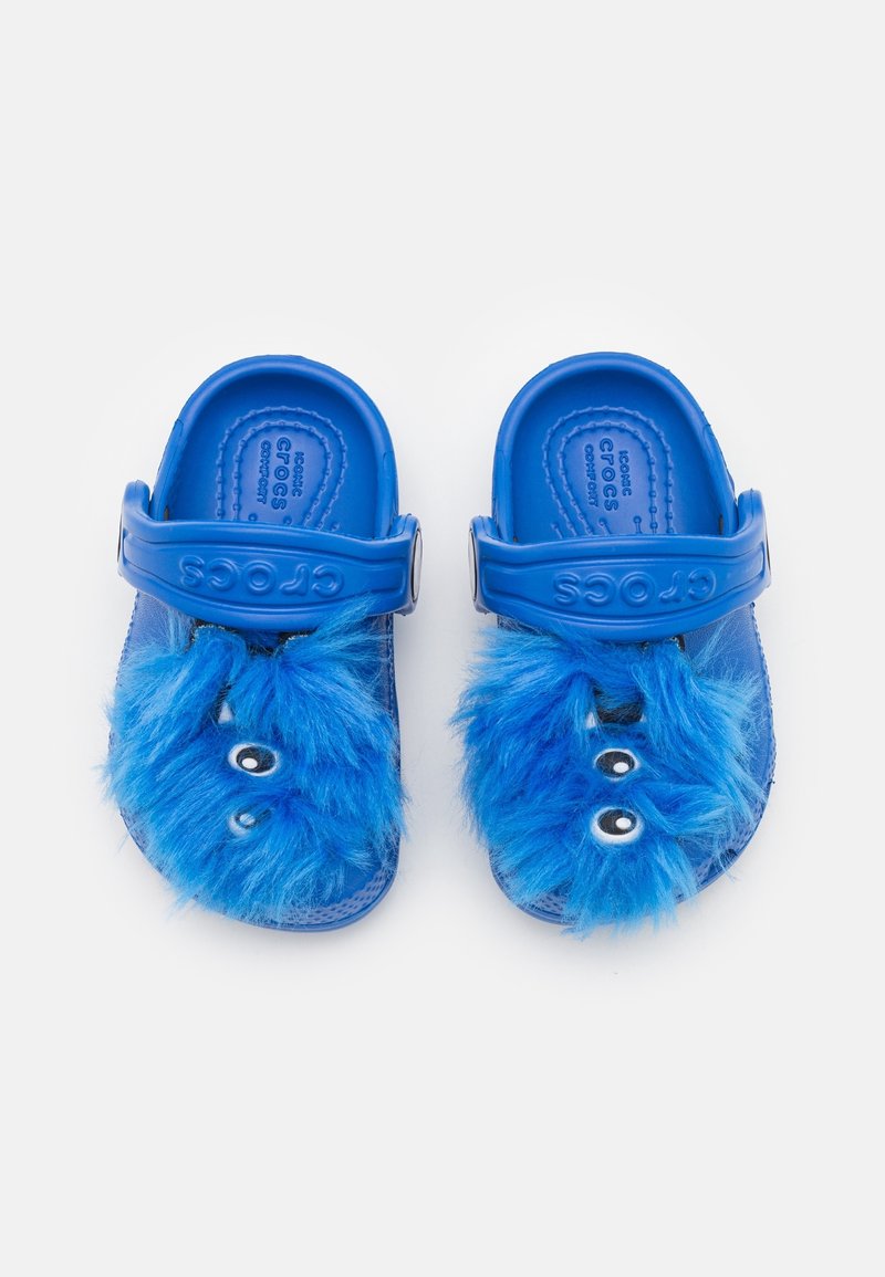 Sandales Crocs bleues pour enfants avec des visages et des yeux de monstre bleus pelucheux sur le dessus, vues de dessus sur fond blanc.