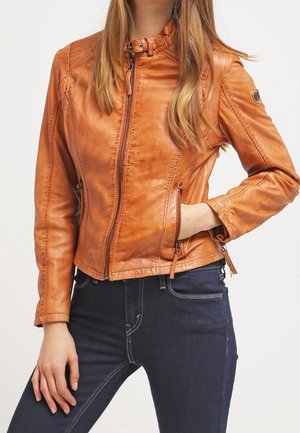 Mujer que lleva una chaqueta ajustada de cuero marrón claro con cremallera y jeans azul oscuro, con una mano en el bolsillo, sobre un fondo claro y sencillo.