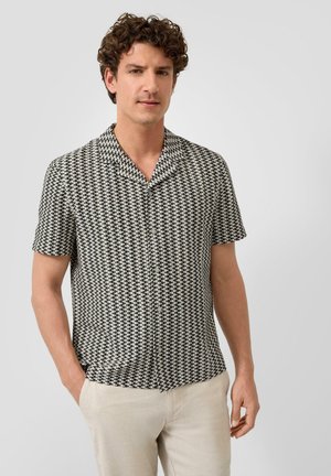 Homme portant une chemise à manches courtes noire et blanche à motifs, boutonnée, avec un pantalon beige, debout devant un fond clair uni.