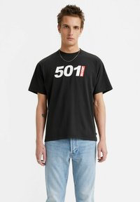 Juodas medvilnės marškinėliai su ryškiu baltu "501" spaudiniu ir raudonu "Levi's" etikete. Trumpomis rankovėmis, laisvo silueto, derinami su šviesiai mėlynomis džinsinėmis kelnėmis.