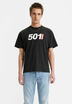 Black cotton t-shirt featuring bold white "501" print and red "Levi's" tag. Short sleeves, relaxed shape, paired with light blue denim jeans.