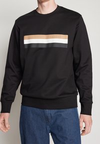 Svart sweatshirt i slät tyg, med en horisontell brun, vit och svart rand samt präglad logotyp över bröstet.