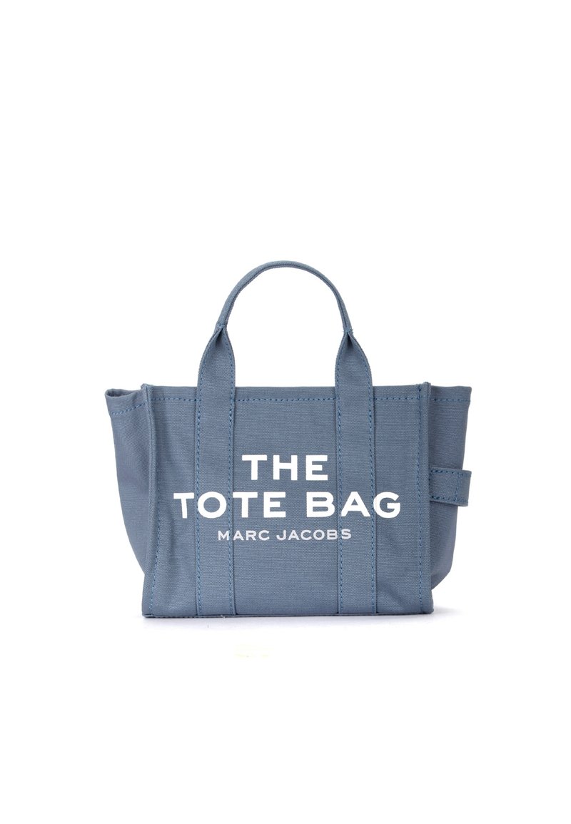 Saco de lona azul com duas alças superiores. Apresenta texto branco: "THE TOTE BAG MARC JACOBS." Design simples e retangular com costuras cosidas.
