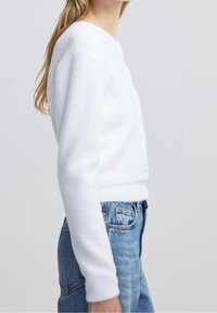 Femme portant un cardigan blanc duveteux à manches longues avec des boutons en perle, associé à un jean en denim bleu taille haute, vue de côté.
