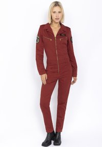 Jumpsuit rosso aderente con colletto a punta, tasche con doppia zip e toppe in stile militare sulle maniche. Il tessuto è liscio e ha una finitura opaca.