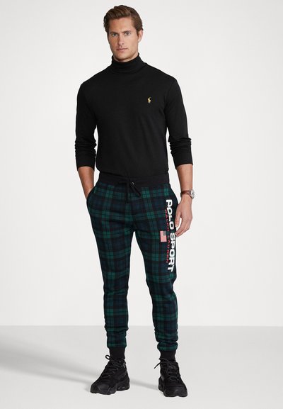Un suéter negro de cuello alto, joggers de cuadros verdes con el logo "POLO SPORT" y zapatillas negras completan el atuendo. Los pantalones tienen puños elásticos.