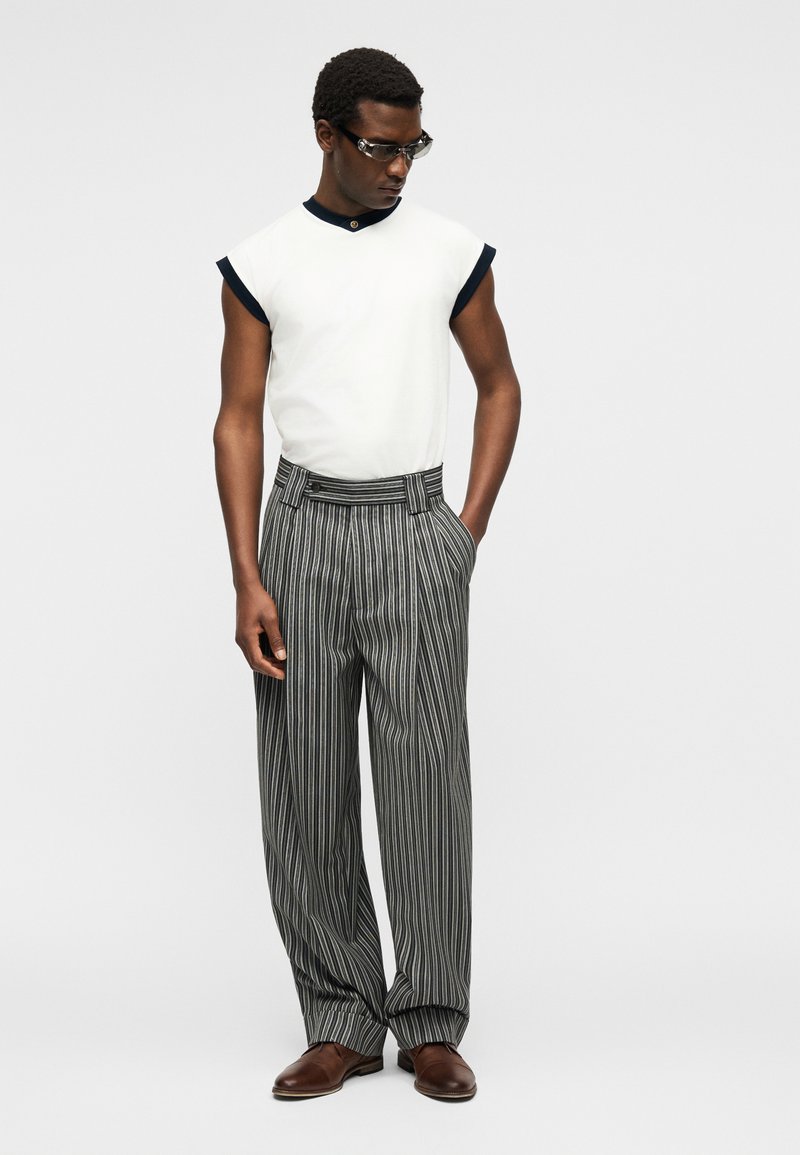 Man wearing sleeveless white top with black trim, gray pinstriped wide-leg pants, brown shoes, and clear wraparound glasses, standing with hands in pockets.