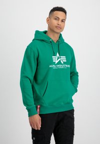 Sudadera verde hecha de una mezcla de algodón con un bolsillo frontal, capucha con cordón y logo blanco. Presenta puños acanalados y una etiqueta en el dobladillo.