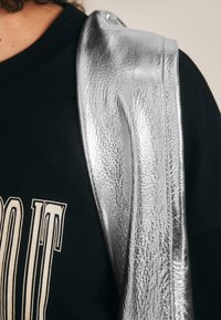 Ezüst színű fémszövet borít egy fekete pólót, amelyen merész, krémszínű felirat található. A szövet felülete sima és fénylő.