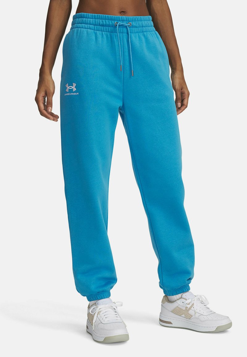 Osoba v jasne modrých teplákach Under Armour s elastickými manžetami, so sťahovacou šnúrkou a bielych teniskách s béžovými akcentmi.