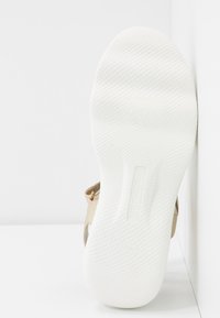 Beige sandál s texturovanou bílou podrážkou, který má pásku s zlatým akcentem a logo výrobce vtlačené do stélky.