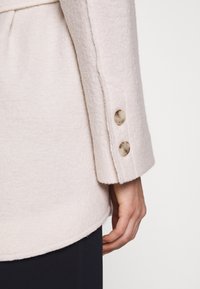 Manche longue d’un manteau en laine écru avec deux boutons beiges sur le poignet, porté sur un pantalon noir, main détendue sur le côté.