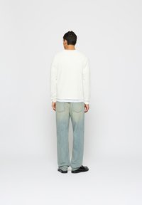 Fehér bordás hosszú ujjú póló kék-zöld átmenetes farmernadrággal, egyenes fazonban. A férfi fekete cipőt visel.