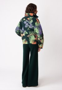 Prevelik temnozeleni flis jopič z barvitimi cvetličnimi motivi, visokim ovratnikom in rebrastimi manšetami; kombiniran z zelenimi širokimi hlačami.