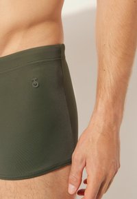 Khaki plavky vyrobené z hladkého, elastického materiálu. Mají přiléhavý střih, ploché švy a malé logo poblíž pásu.
