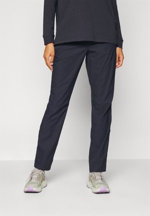 Person iført sorte bukser med lige ben og lysegrå sneakers med lilla såler, stående mod en ensfarvet lys baggrund.