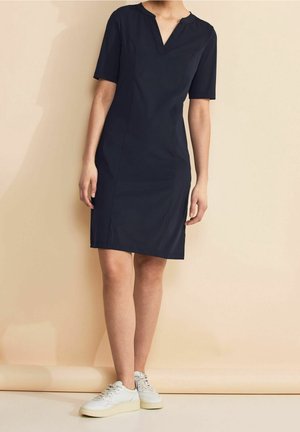 Femme portant une robe bleu marine mi-longue avec manches courtes et col en V, associée à des baskets blanches, debout devant un fond beige.