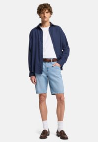 Granatowa koszula z guzikami noszona na białej koszulce, w zestawie z jasnoniebieskimi szortami dżinsowymi. Brązowy pasek, białe skarpetki i ciemne buty wsuwane. Kręcone włosy.