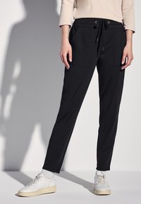 Pantalones de chándal negros con cinturilla ajustable, dos bolsillos frontales y un diseño ajustado, combinados con zapatillas deportivas blancas y calcetines grises.