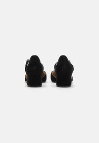 Chaussures à talons en suede noir avec un dos fermé et des côtés ouverts, présentant un talon épais et un détail de sangle ajustable. Texture lisse et design élégant.