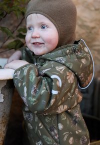 Zielona dziecięca kurtka z nadrukami o tematyce przyrodniczej, wyposażona w kaptur, zapinana na zamek błyskawiczny i posiadająca miękką podszewkę. Dziecko ma na głowie brązową dzianą czapkę.