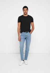 Czarny t-shirt z krótkim rękawem, jasnoniebieskie dżinsy, białe trampki. Codzienny strój o luźnym kroju z okrągłym dekoltem i prostym designem, na tle białej ściany.