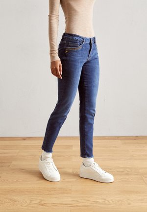 Person, die eng anliegende blaue Jeans mit Reißverschluss- und Nieten-Details, beige langärmliges Oberteil, weiße Socken und weiße Turnschuhe trägt und auf einem Holzboden steht.