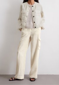 Lys beige linned jakke med sorte knapper, parret med løstsiddende beige cargobukser med sidelommer og flade sorte sandaler.