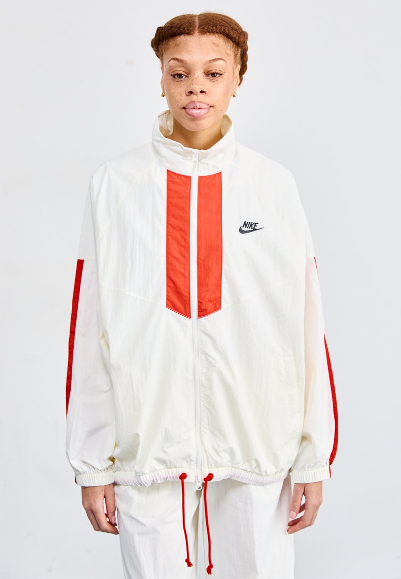 Fehér szélálló dzseki piros panel részletekkel, zipzárral, elasztikus mandzsettákkal és húzózsinóros derékkal. Nike logó a mellkason.