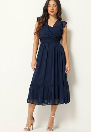 Femme portant une robe midi bleu marine sans manches avec un ourlet volanté et des sandales argentées à talons hauts, debout devant un fond blanc.