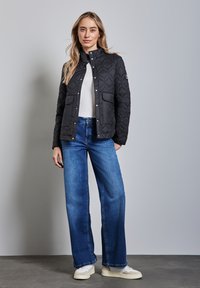 Schwarze gesteppte Jacke mit Druckknöpfen, zwei vorderen Taschen, getragen über einem weißen Oberteil und blauen weiten Jeans, ergänzt durch hellfarbige Sneaker.