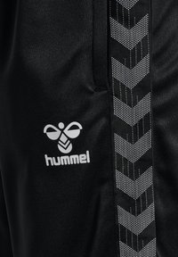 Tissu noir avec le logo blanc Hummel et une bande verticale à motif chevron sur le côté, probablement une partie de vêtements de sport ou de vêtements d’athlétisme.