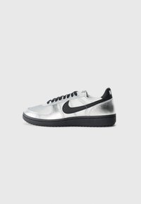 Strieborná a čierna nízka teniska Nike s čiernym swooshom, čiernymi šnúrkami a čiernou podrážkou na jednoduchej svetlošedej ploche.
