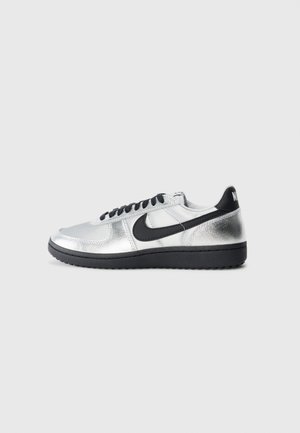 Sneaker Nike low-top argento e nero con swoosh nero, lacci neri e suola nera su uno sfondo grigio chiaro semplice.