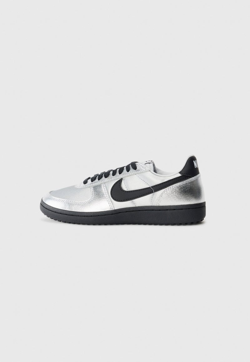 Strieborná a čierna nízka teniska Nike s čiernym swooshom, čiernymi šnúrkami a čiernou podrážkou na jednoduchej svetlošedej ploche.