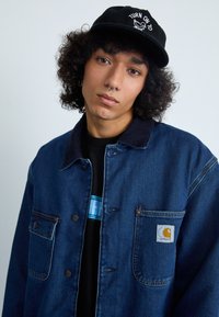 Traper jakna s tamnim pranjem, prednjim džepovima i Carhartt logom. Crna kapa s natpisom "TURN ON TO". Crna majica ispod.