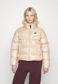Šviesiai rožinė, paminkštinta, trumpa striukė su aukštu apykakle, priekyje užtrauktuku ir juodu Nike logotipu ant krūtinės. Lygus, blizgus paviršius.