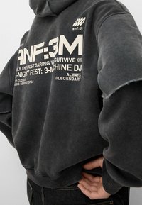 Sudadera negra de gran tamaño con dobladillo desgastado, con un gran texto gráfico blanco en la espalda. Fabricada con un tejido suave y texturizado.