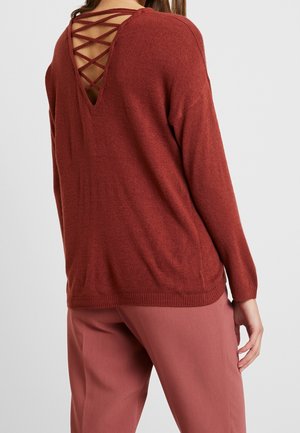 Kobieta ubrana w sweter w kolorze rdzawej cegły z głębokim dekoltem V na plecach i paskami skrzyżowanymi na krzyż, w połączeniu z różowymi spodniami w kolorze pudrowego różu, stojąca na jednolitym tle.