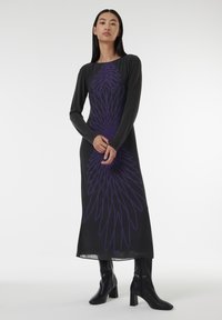 Robe noire longue avec un motif violet ressemblant à des plumes, manches longues et silhouette droite, associée à des bottes à talons noires.