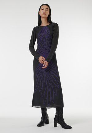 Robe noire longue avec un motif violet ressemblant à des plumes, manches longues et silhouette droite, associée à des bottes à talons noires.