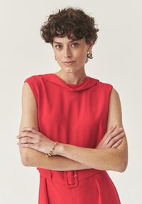 Frau mit kurzen lockigen Haaren, die ein ärmelloses rotes Kleid, ein goldenes Armband und Ohrringe trägt, stehend mit verschränkten Armen vor einem schlichten Hintergrund.