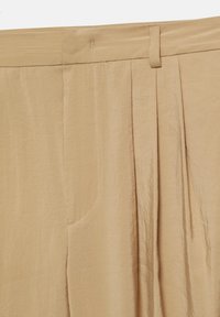 Beige plisserede bukser med båndlinning, bæltestrop og synlige sømme på tekstureret stof.