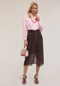 Camicia rosa a maniche lunghe con tasche frontali, gonna avvolgente marrone con nodo drappeggiato, scarpe con cinturini nere, e una piccola borsa a due toni.